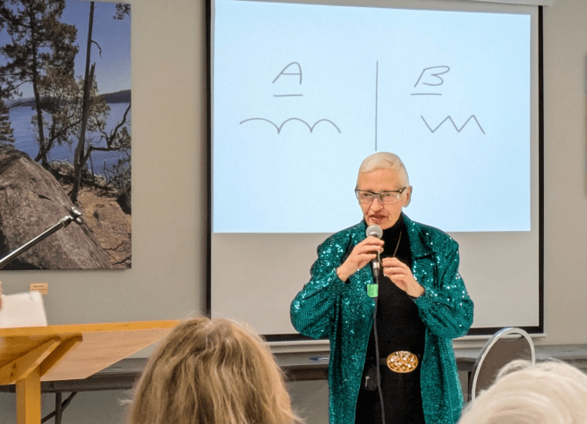 Presenter in a green sequined jacket speaking before a screen showing a diagram labeled A with a wavy line and B with a zigzag line.